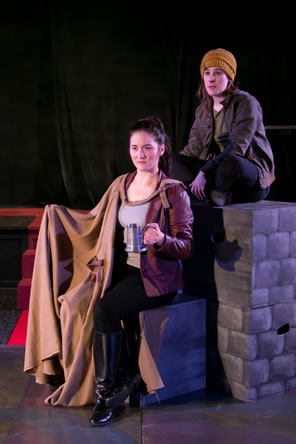 Hallie in red leather jacket, t-shirt, black pants and black boots with a brown cloak and holding a tankard sits on a block while Peto in yellow stocking cap, brown jacket and black jeans sits cross-legged on a stack of stone-wall painted blocks